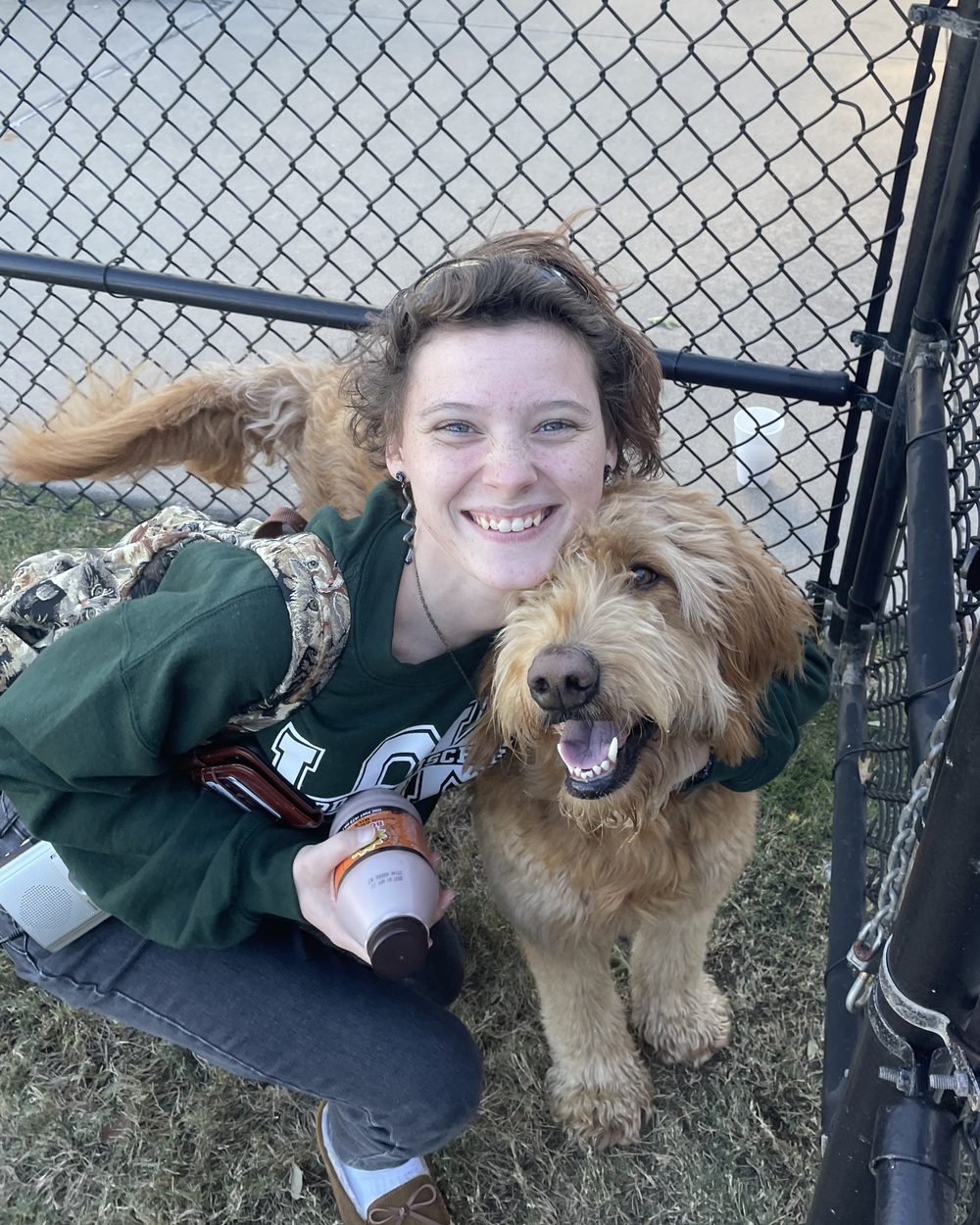 Abby crouching beside a happy goldendoodle in a fenced yard.